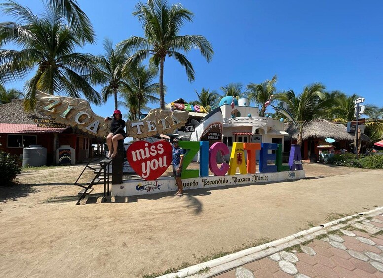Un letrero colorido con las palabras 'Miss you' y 'Ticotela' en un entorno playero, con palmeras y un edificio de techo de paja. 2