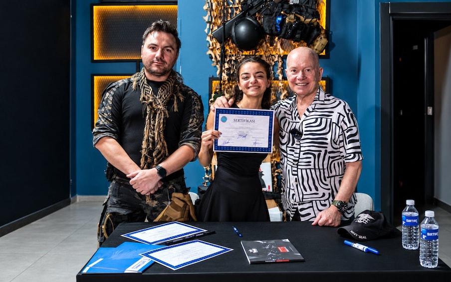 Three people standing at a table with a certificate, a black bag, and water bottles. 4