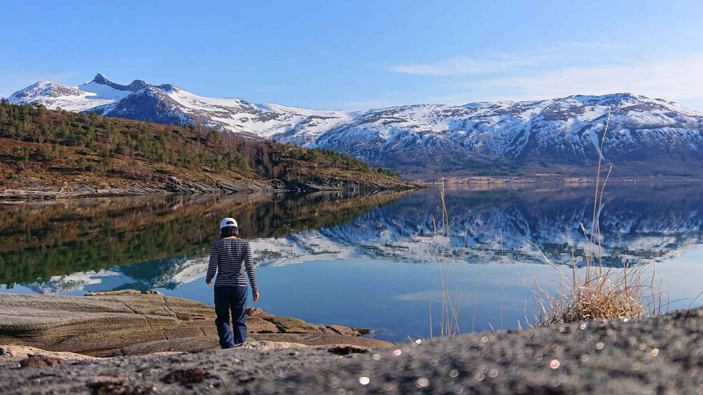 Une personne se tient sur une côte rocheuse, surplombant un lac paisible, avec des montagnes enneigées en arrière-plan. 4