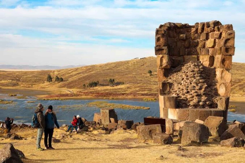 A stone ruin with people standing nearby, a river in the background, and a hilly landscape. 5