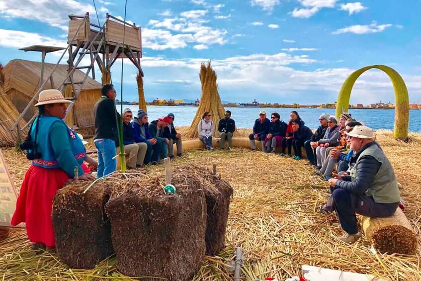 A group of people sitting on a straw-covered ground, with a body of water and a wooden structure in the background. 2