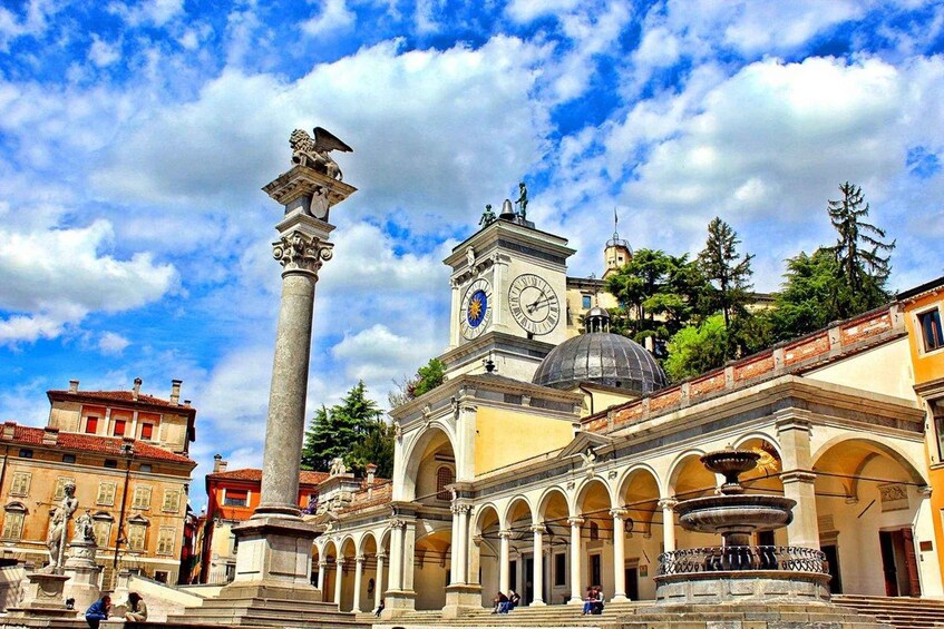 A historic square with a prominent column, a clock tower, and a fountain. 1