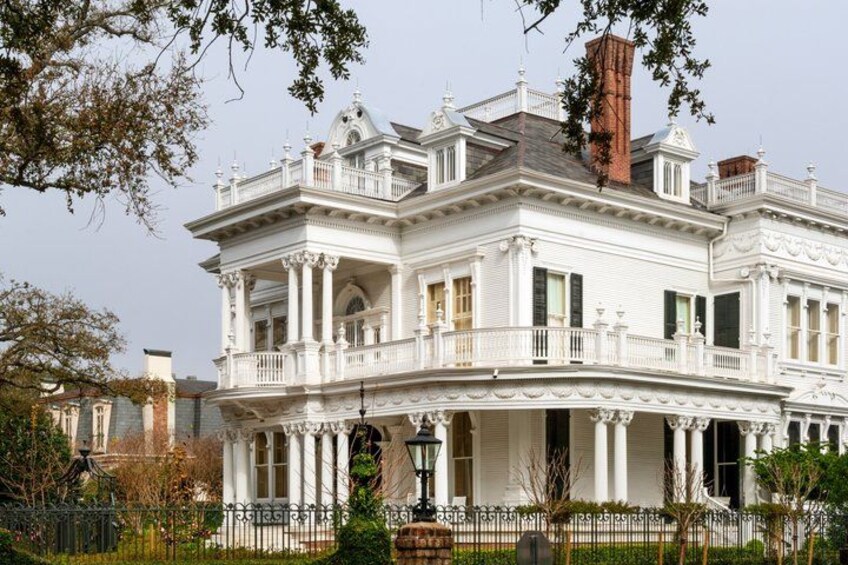 A large white mansion with multiple balconies and columns, surrounded by a fence and trees. 2