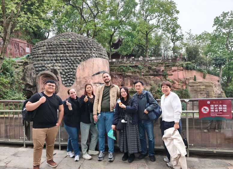 A group of people posing in front of a large Buddha statue with a rocky background and trees. 1