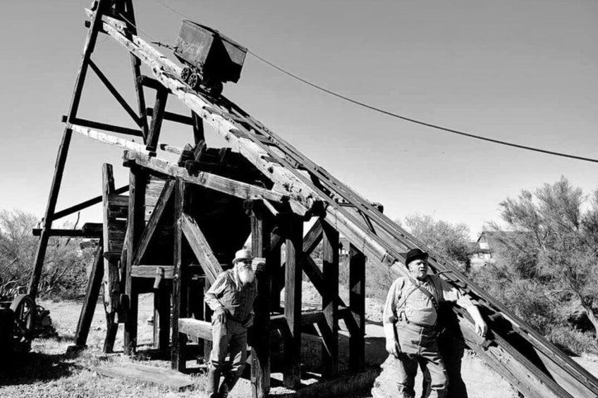 A dilapidated wooden structure with a large mechanical component, possibly a pump or engine, and two individuals standing nearby. 3