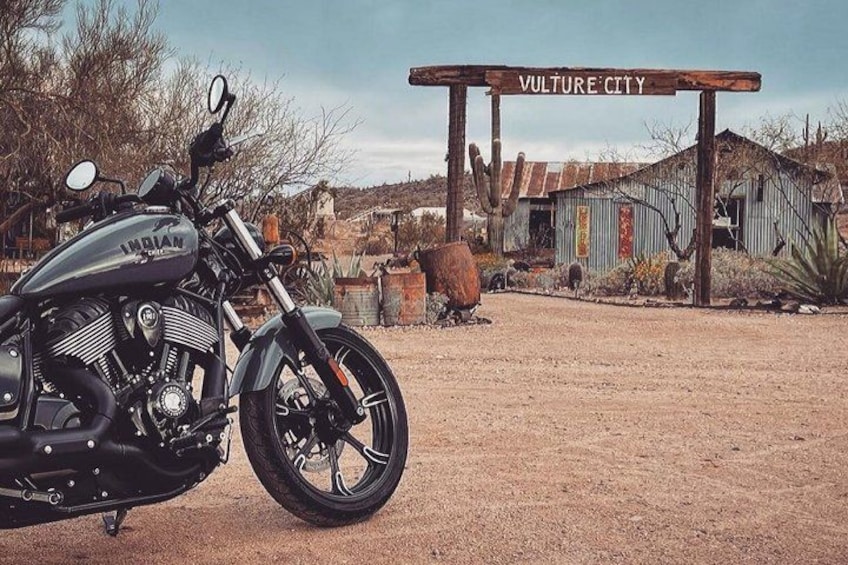 A vintage motorcycle parked in front of a rustic building labeled 'Vulture City.' 1