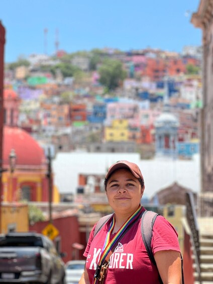 Una persona con una camisa roja y un lanyard con arcoíris, de pie frente a una ladera colorida con edificios. 1