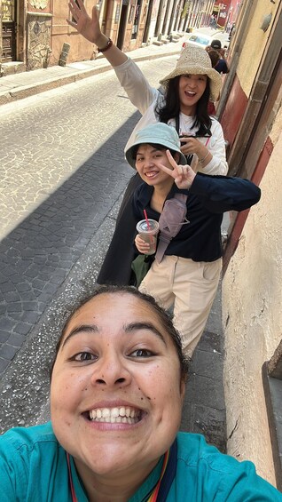 Dos personas tomándose un selfie en una calle adoquinada, con edificios y un coche al fondo. 2