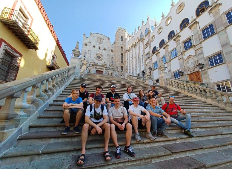 Un grupo de personas sentadas en escalones de piedra frente a un edificio histórico con arquitectura ornamentada. 4