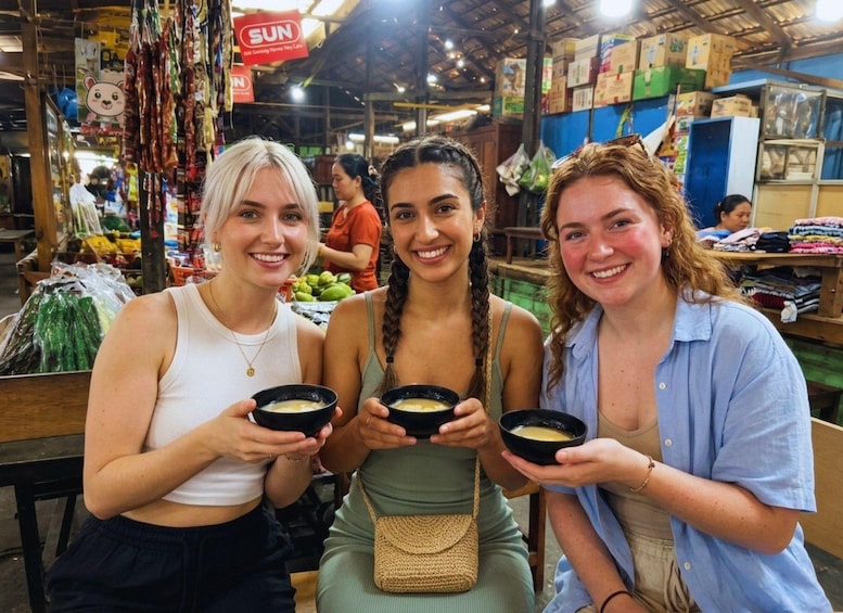 Three people holding bowls of food in a market with various goods and a 'SUN' sign in the background. 2