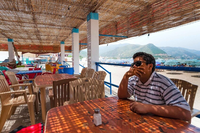 Un hombre sentado a una mesa con mantel estampado, bajo una estructura de techo tejido, con vista a una playa con sombrillas y montañas al fondo. 3