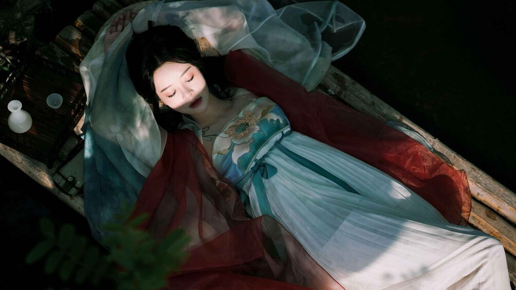 A person lying in a boat, wearing a traditional dress with a red shawl, and a white head covering. 1