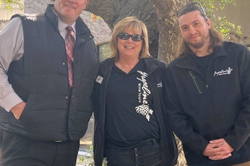 Three people standing outdoors, one wearing a vest, another in a black shirt with a wine logo, and the third in a black jacket. 2
