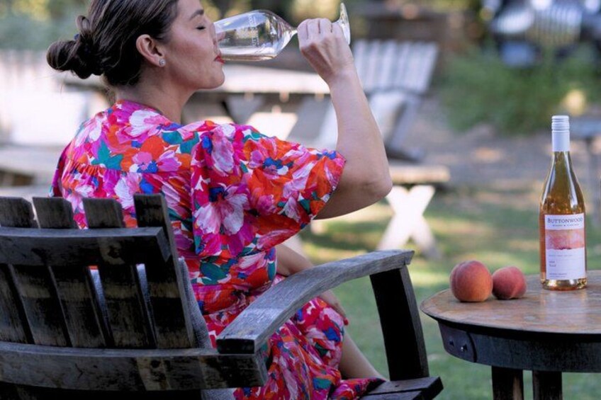 A person sitting on a wooden bench outdoors, wearing a floral dress, with a bottle of wine and peaches on a table beside them. 5