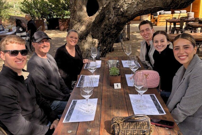A group of people seated at a wooden table outdoors with wine glasses and papers. 3