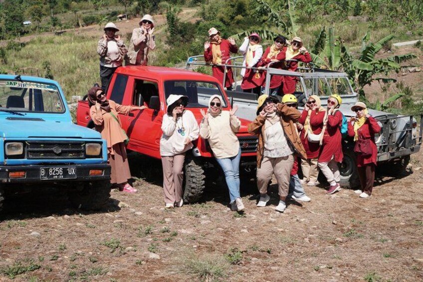 A group of people in outdoor attire near a red pickup truck and a blue car, with a dirt ground and vegetation in the background. 2