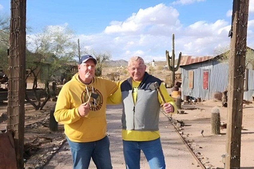 Two people standing outdoors with cacti and a wooden structure in the background. 3