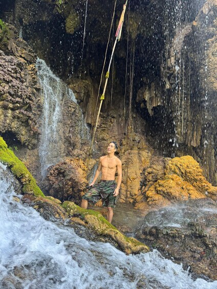 Una persona de pie cerca de una cascada en una zona rocosa con rocas cubiertas de musgo y una cuerda colgando de un árbol. 2