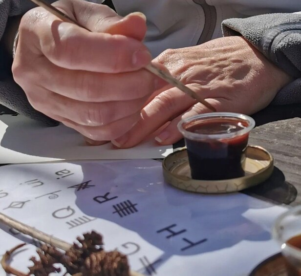 A person writing on paper with a brush, a small cup of ink, and a pine cone on the table. 3