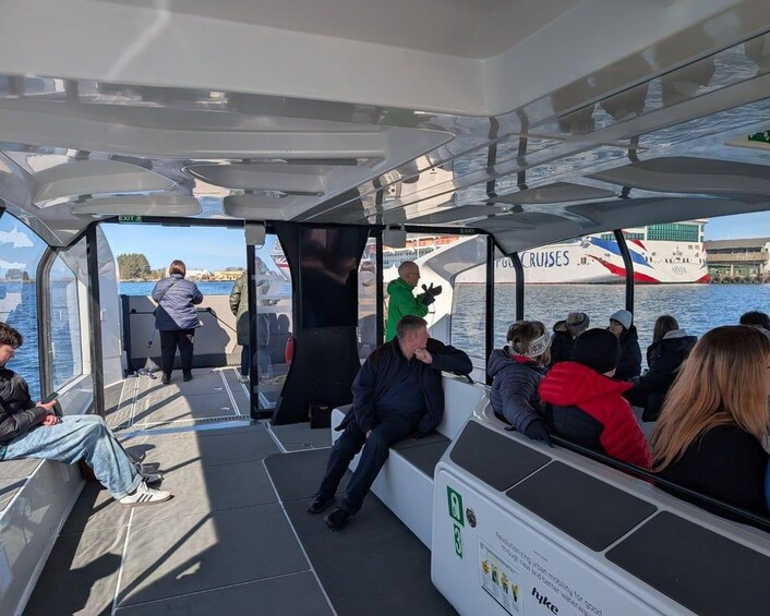 A ferry with passengers seated and standing, a person in a green jacket walking, and a large cruise ship in the background. 4