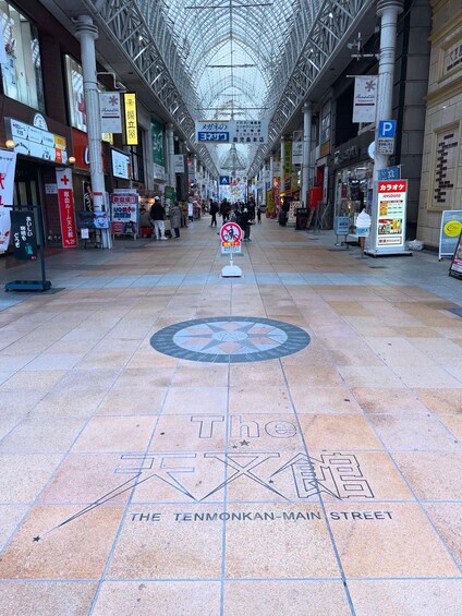 A pedestrian walkway with a tiled floor, overhead metal structure, and various signs and advertisements. 2