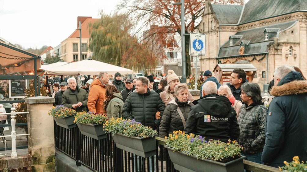 Ryhmä ihmisiä oli kokoontunut katumarkkinoiden ympärille. Markkinapaikalla oli kukkaistutuksia, ja taustalla näkyi historiallinen rakennus. 2