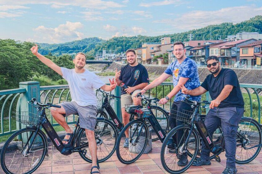 Four people on a bridge with bicycles, greenery in the background, and buildings. 1