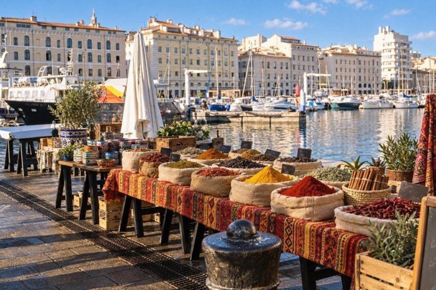 A seaside market with tables displaying spices and herbs, boats docked in the background, and buildings along the waterfront. 5