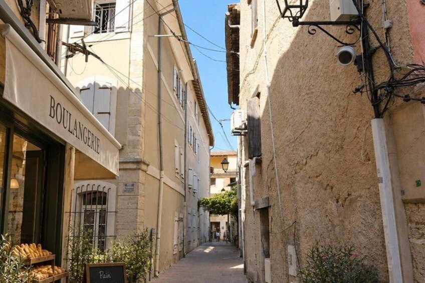 A narrow street with buildings on either side, a bakery with bread displayed outside, and a sign that reads 'BOULANGERIE'. 2
