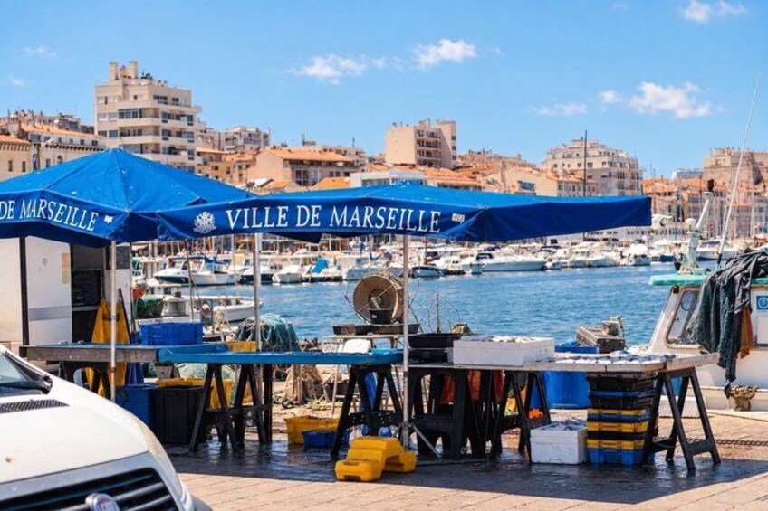 A seaside market with blue umbrellas labeled 'Ville de Marseille,' boats docked in the background, and a white car in the foreground. 1