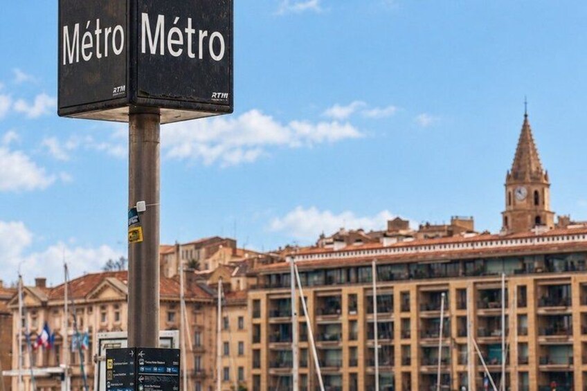 A metro sign with 'Metro' written in two languages, a clock, and a building with a tower in the background. 3