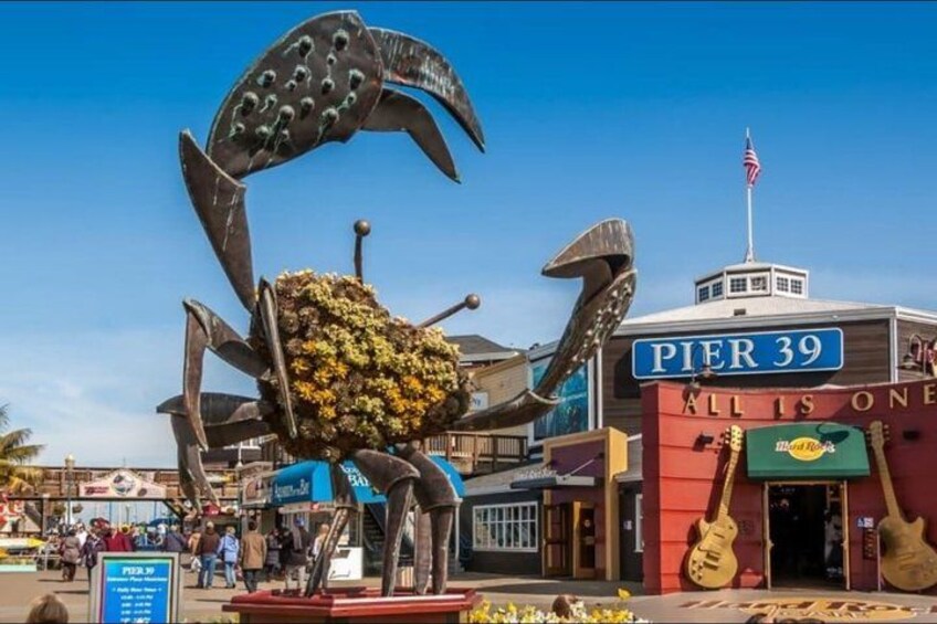 A large crab sculpture in front of Pier 39, with a sign reading 'All is One' and a guitar-shaped building. 3