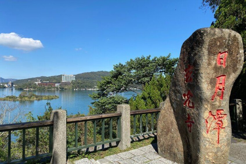 A stone monument with red inscriptions, a wooden fence, and a view of a lake with buildings in the background. 4