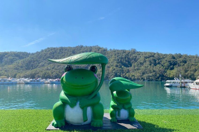Two green dinosaur statues on a grassy area by a lake with boats and a forested hill in the background. 3