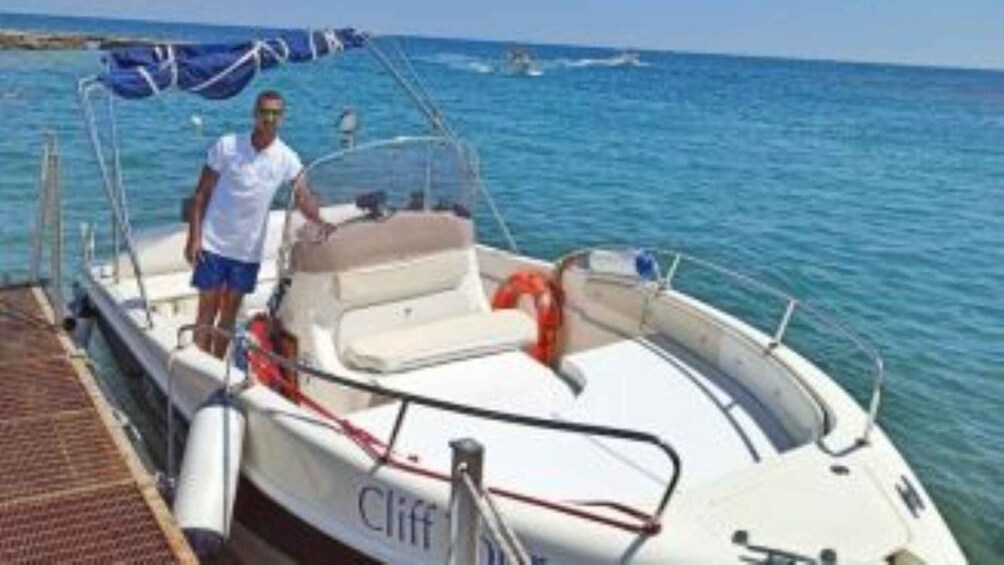 Ein kleines, weißes Boot mit einer Person, die am Steg steht, einem Rettungsring und einem weiteren Boot im Hintergrund. 1