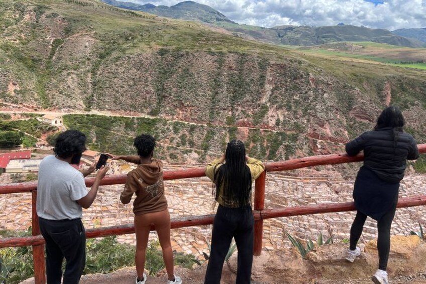 Four people standing on a ledge, overlooking a mountainous landscape with a river and buildings in the valley below. 3