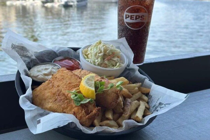 A meal of fish and chips, coleslaw, and a drink on a table by a body of water. 5