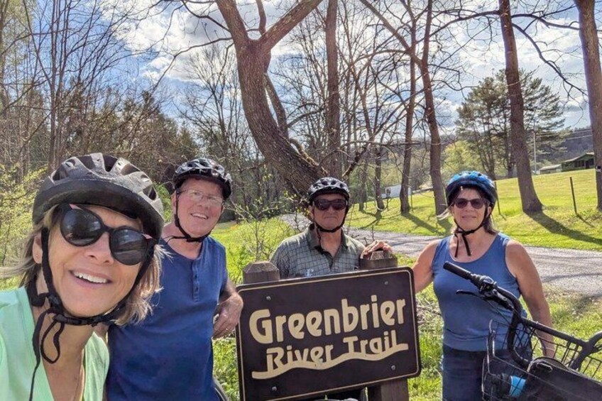Four people with bicycles, a sign for the Greenbrier River Trail, and a rural landscape with trees and a clear sky. 3
