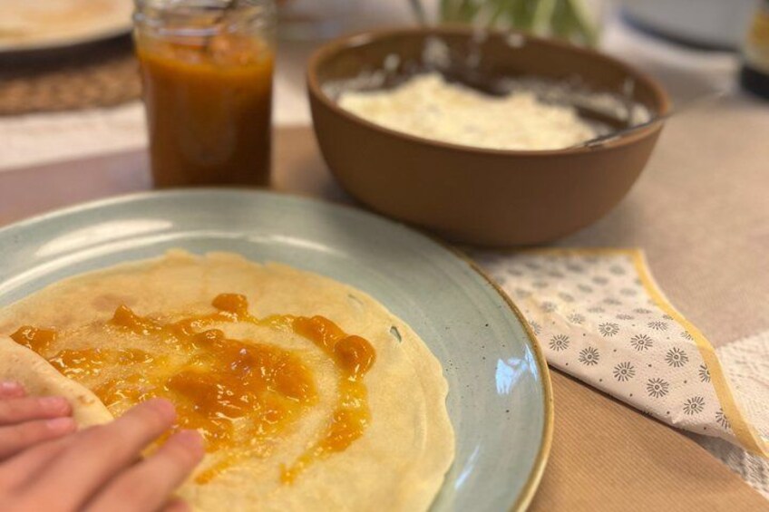 A crepe being spread with a yellow sauce on a plate, with a bowl of batter and a jar of sauce in the background. 3