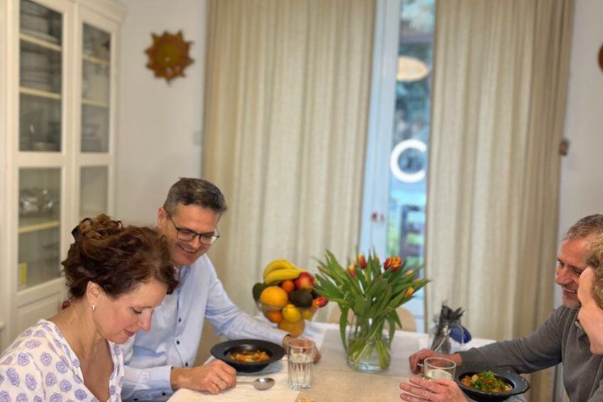 A dining table with people seated, a vase with flowers, and a fruit bowl. 1