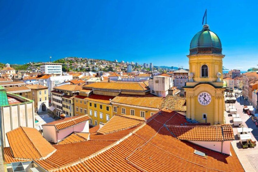 A cityscape with a prominent clock tower, terracotta-roofed buildings, and a clear blue sky. 1