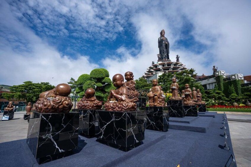 A bronze statue of a seated figure, surrounded by smaller bronze figures, in a public square with a clear sky. 3