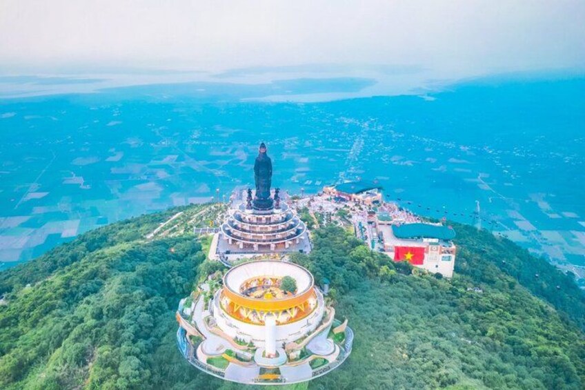 A large statue on a hilltop with a circular building below, surrounded by greenery and a coastal view. 1
