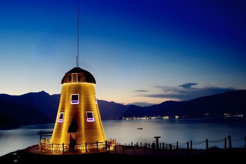 A lighthouse with a unique conical shape, a fence, and a calm lake with mountains in the background. 2