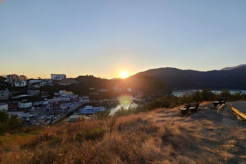 A sunset over a coastal town with buildings, a marina, and benches on a hillside. 4