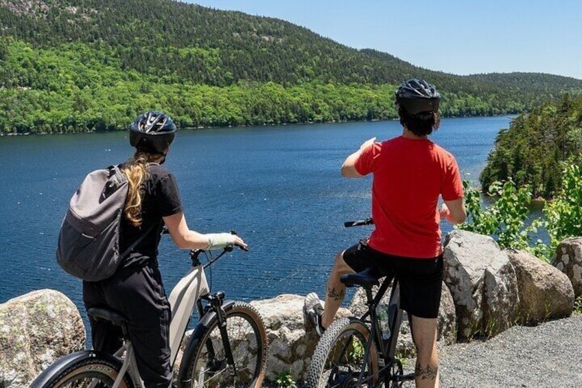 Two cyclists on a rocky path overlooking a lake with forested hills in the background. 4