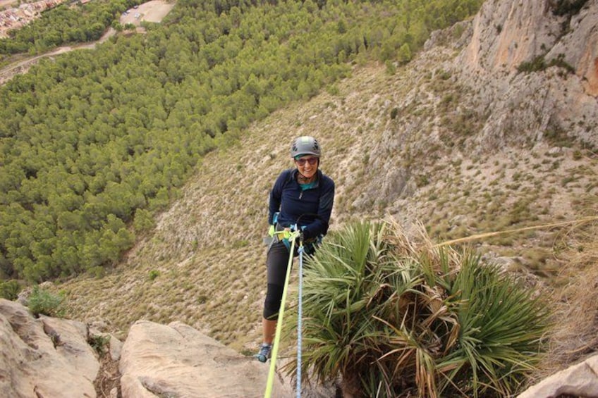 A person rappelling down a rocky hillside with a rope, surrounded by dense green forest and rocky terrain. 3