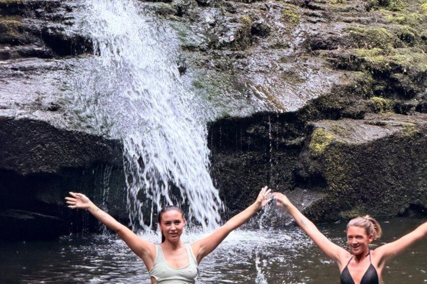 Two people standing in water with arms raised, a waterfall in the background. 2