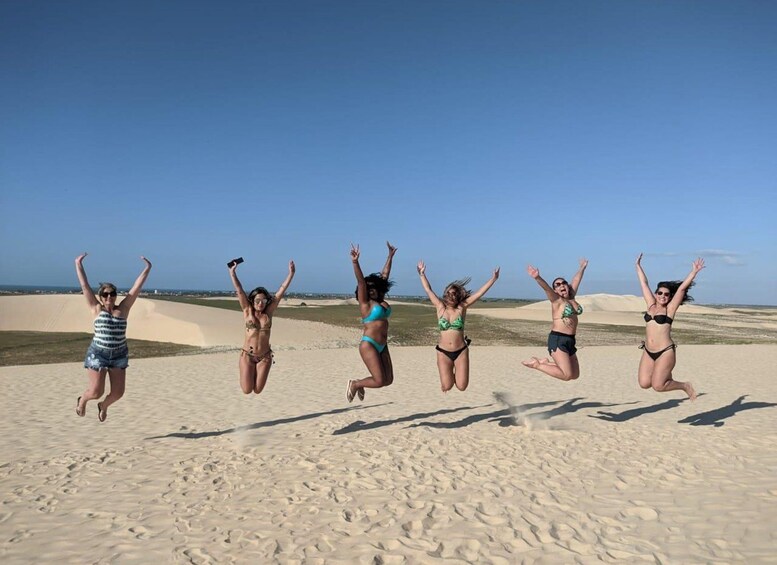 Cinco personas saltando en una playa arenosa con dunas al fondo. 4