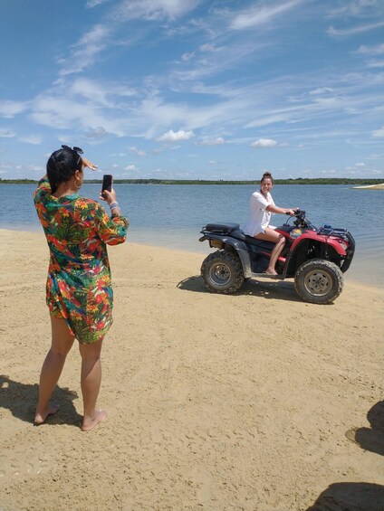 Una persona fotografiando a otra persona sobre un vehículo todo terreno rojo en una playa arenosa, con un lago al fondo. 1